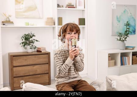 Kleiner Junge mit Mikrofon und Kopfhörern, der zu Hause auf dem Sofa singt Stockfoto