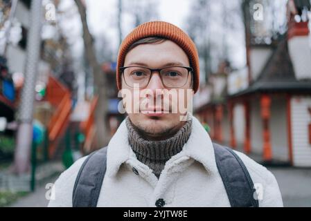 Ein selbstbewusster Mann mit orangefarbener Mütze, Brille und weißer Sherpa-Jacke steht in einem Vergnügungspark und strahlt Stil und Individualität aus Stockfoto