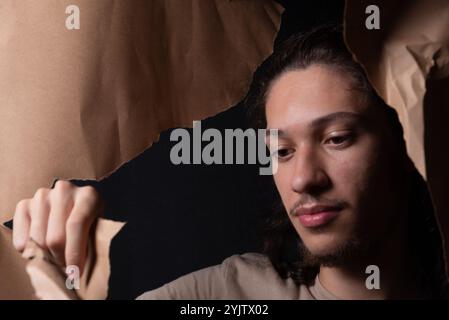 Ein junger Mann, der durch ein Loch in einem Blatt braunem Papier rauskommt. Nahaufnahme. Studio-Porträt. Stockfoto