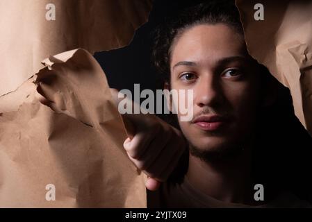 Ein junger Mann, der durch ein Loch in einem Blatt braunem Papier rauskommt. Nahaufnahme. Studio-Porträt. Stockfoto