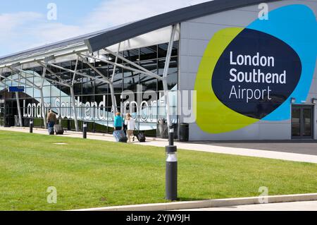 Flughafen London Southend. Passagiere mit Gepäck, die in Richtung des Terminalgebäudes fahren, mit dem London Southend Airport Logo und dem Symbol Stockfoto