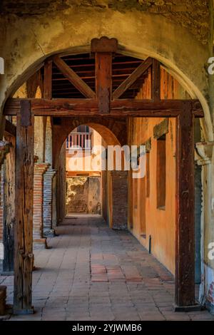 San Juan Capistrano, USA – 6. August 2023: Verwitterte Holzbalken und Bogengänge umrahmen einen sonnendurchfluteten Durchgang bei Mission San Juan Capistrano Stockfoto