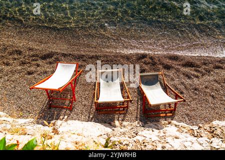 Drei leere Liegen an einem Kieselstrand mit Blick auf das ruhige, klare Wasser des Ozeans an einem sonnigen Tag. Stockfoto