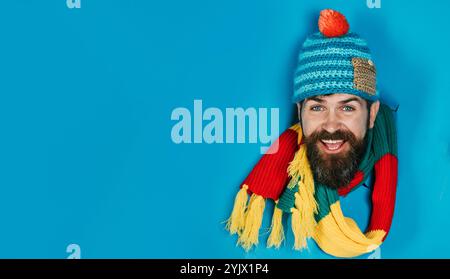 Lächelnder bärtiger Mann, der durch ein Loch in Papier schaut. Hübscher Mann in Strickmütze und farbenfrohem Schal. Warme Kleidung für Herren. Herren Herbst-Winter-STYLE. Meer Stockfoto