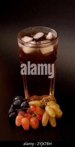 Traubensaft mit Eis in einem hohen Glas auf schwarzem Hintergrund, Zweige verschiedener Obstsorten in der Nähe. Nahaufnahme, vertikale Anordnung. Stockfoto