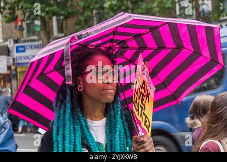 London, Großbritannien. Juli 2017. Eine Frau mit einem rosa gestreiften Regenschirm hält ein Black Lives Matter-Plakat vor der Stoke Newington Polizeistation für Rashan Charles, der starb, als zwei Polizisten ihn mit Handschellen in einem Geschäft an der Kingsland Rd in den frühen Morgenstunden des Samstag, den 22. Juli, auf dem Boden festhielten. Seine Familie ist entschlossen, Antworten auf seinen Tod zu bekommen, aber sie fordert alle auf, innerhalb des Gesetzes zu handeln. Mitglieder der Familie Charles kamen zusammen mit der Familie Edson da Costa zu dem Protest, der nach der Verhaftung in East Ham im Juni starb. Die Demonstranten hielten Bilder der beiden Männer und Darren Cumberb Stockfoto
