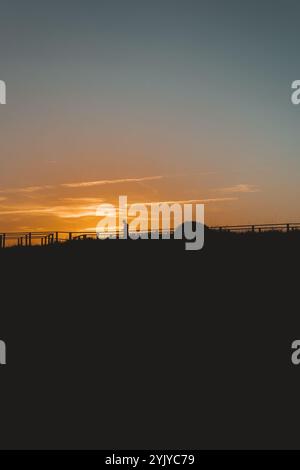 Eine einsame Figur mit Silhouetten läuft entlang einer Promenade vor einem leuchtenden Sonnenunterganghimmel, mit viel negativem Platz am unteren Rand für Text Stockfoto