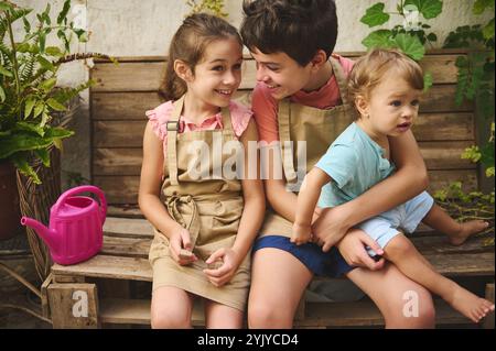 Drei fröhliche Kinder in lässiger Kleidung, die einen Tag im Garten verbringen. Sie sitzen auf einer rustikalen Holzbank, interagieren spielerisch, umgeben von B Stockfoto