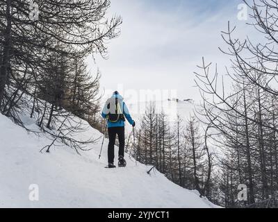 Ein einsamer Wanderer in Schneeschuhen durchquert einen verschneiten Bergweg, der von Bäumen und zerklüfteten Gipfeln umgeben ist, und genießt ein friedliches Winterabenteuer in der Natur. Stockfoto