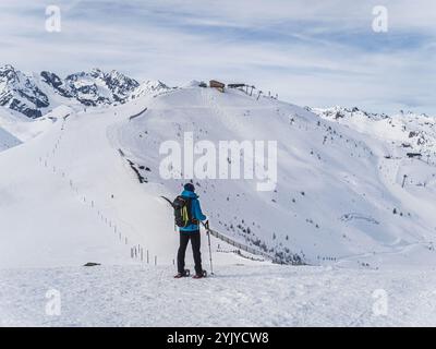 Ein einsamer Wanderer in Schneeschuhen durchquert einen verschneiten Bergweg, der von Bäumen und zerklüfteten Gipfeln umgeben ist, und genießt ein friedliches Winterabenteuer in der Natur. Stockfoto