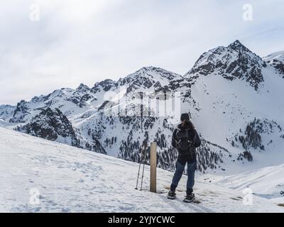 Ein einsamer Wanderer in Schneeschuhen durchquert einen verschneiten Bergweg, der von Bäumen und zerklüfteten Gipfeln umgeben ist, und genießt ein friedliches Winterabenteuer in der Natur. Stockfoto