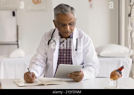 Doktor sitzt am Schreibtisch, hält Tablette und schreibt in Tagebuch Stockfoto