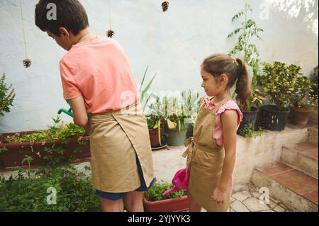 Kleine Kinder, die Schürzen tragen, neigen dazu, im Garten zu Pflanzen. Der Junge tränkt, während das Mädchen beobachtet, und lernt etwas über Gartenarbeit und Natur. Stockfoto