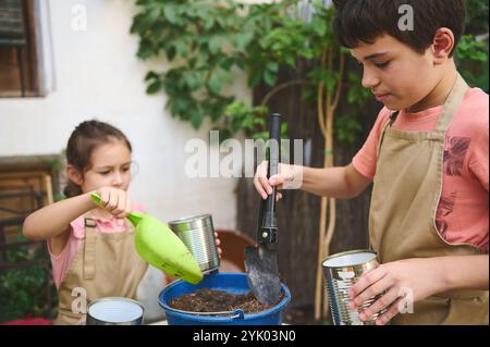 Zwei Kinder genießen Gartenarbeit, indem sie recycelte Blechdosen und Erde zum Pflanzen von Samen verwenden. Sie nehmen an einer Bildungsaktivität im Freien Teil und fördern die Teamarbeit Stockfoto