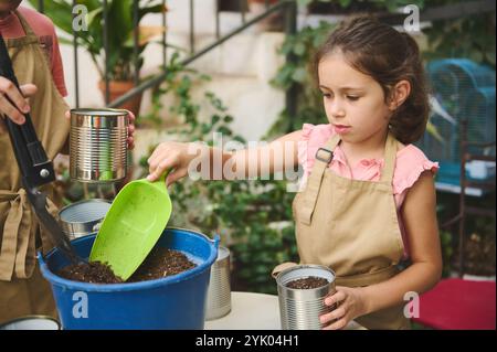 Ein junges Mädchen, das eine Schürze trägt, im Garten arbeitet und recycelte Blechdosen mit Erde füllt. Bei einer Pflanzaktion im Freien demonstriert sie die Umwelt Stockfoto