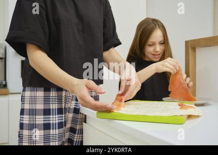 Die Tochter hilft ihrer Mutter, die geräucherte Forelle auf das Fladenbrot zu legen. Das Konzept des gemeinsamen Kochens zu Hause. Stockfoto