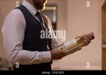 Schuss eines nicht erkennbaren männlichen Sommeliers in Stripphemd und Indigo-Anzug-Weste, der eine Flasche Weißwein hält Stockfoto