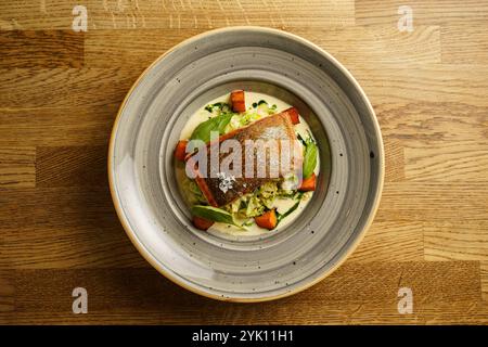 Ein perfekt gegrilltes Lachsfilet, serviert mit geröstetem Gemüse und einer cremigen Sauce auf einem dekorativen Teller. Das Gericht ist mit Spinatblättern garniert, Stockfoto