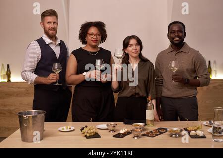 Kruppe Porträt multirassischer professioneller Sommeliers, die Glas mit Weißwein halten und lächelnd, Holztisch mit einem Snack für Wein vor ihnen Stockfoto