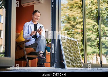 Glückliche Frau mit Smartphone, das mit Photovoltaik-Solarmodul verbunden ist. Integration nachhaltiger erneuerbarer Energien in den Alltag, demonstrieren die praktische Nutzung von Solarstrom für Ladegeräte. Stockfoto