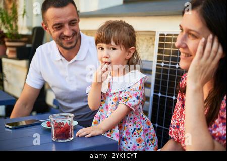 Glückliche Familie, die Zeit zusammen am sonnigen Tag verbringt. Ein kleines Mädchen lächelt ihren Eltern zu. Ein junges Paar und ihr Kind sitzen draußen auf dem Hintergrund eines Solarmoduls. Stockfoto