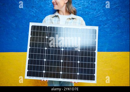 Frau, die Solarpanel zum Laden hält. Die lächelnde Frau trägt ein kariertes Hemd über weißem Oberteil und steht vor leuchtend blauem und gelbem Hintergrund der ukrainischen Flagge. Stockfoto