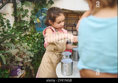 Ein junges Mädchen auf einer Schürze pflanzt Samen in recycelten Blechdosen in einem üppigen Garten und fördert so Umweltfreundlichkeit und Kreativität. Stockfoto