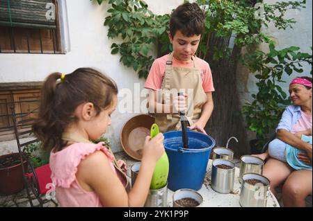 Zwei Kinder arbeiten im Garten, indem sie Boden in Töpfen mischen, während ein Erwachsener im Freien beaufsichtigt. Die Szene strahlt Teamarbeit und Lernen aus. Stockfoto