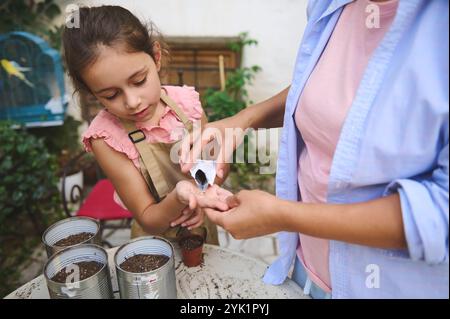 Eine liebevolle Mutter hilft ihrer Tochter, Samen in kleinen Töpfen zu Pflanzen. Das Mädchen trägt eine Schürze und konzentriert sich auf die Aufgabe. Gartenwerkzeuge und Töpfe sind sichtbar Stockfoto