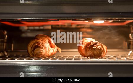 Zwei Croissants, die in einem Ofen mit warmem Licht backen, beleuchten das goldblättrige Gebäck während des Kochvorgangs Stockfoto