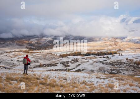 Ein einsamer Fotograf in roter Jacke fängt die schneebedeckten Berge des Altai ein, umgeben von einer dramatischen Winterlandschaft mit sanften Hügeln und bewölktem Himmel. Stockfoto