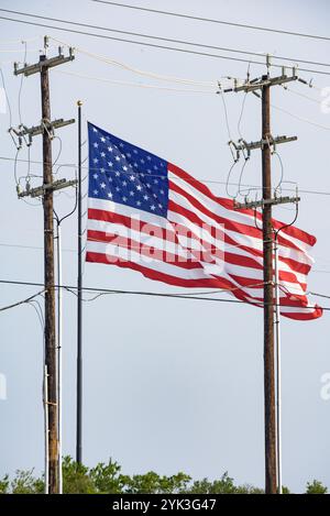 Die US-Flagge winkt am 5. Juni 2024 hinter Strommasten im Bexar County, Texas. Die Bereitstellung zuverlässiger und erschwinglicher Elektrizität ist von entscheidender Bedeutung, um das wirtschaftliche Wohlergehen und die Lebensqualität aller Landbewohner des Landes zu erhalten. Die USA Landwirtschaftsministerium (USDA) Rural Development (RD) Rural Utilities Service (RUS) Electric Program bietet Führungsrolle und Kapital, um Amerikas riesige ländliche elektrische Infrastruktur zu erhalten, zu erweitern, zu modernisieren und zu erweitern. Unter der Autorität des Rural Electrification Act von 1936, das Electric Program, macht die Elektrizitätswerke der USA Stockfoto