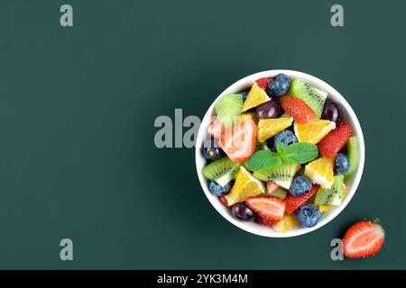 Obst- und Beerensalat auf grünem Hintergrund. Obstsalat aus Erdbeeren, Orange, Kiwi und Trauben in einer weißen Schüssel. Stockfoto