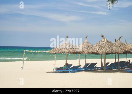 Strohschirme und Sonnenliegen am leeren Kiesstrand mit Meer im Hintergrund. Einsamer Strand mit Rattan-Liegestühlen und Sonnenschirmen Stockfoto