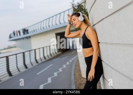 Eine junge Frau in sportlicher Kleidung lehnt sich mit Kopfhörern an eine schlanke Wand und nimmt sich nach dem Joggen in einer lebhaften städtischen Umgebung einen Moment Zeit, um ihre Musik zu genießen Stockfoto