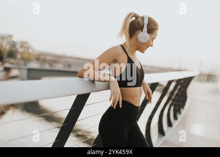 In sportlicher Ausrüstung hält eine junge Frau während ihres Joggs an, um Musik zu genießen, und lehnt sich an ein Geländer mit einer friedlichen Kulisse aus Wasser und Stadtleben Stockfoto