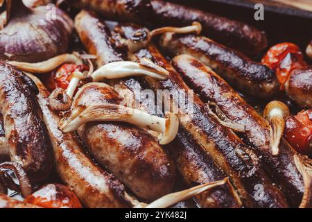 Grillpfanne mit leckeren hausgemachten Hühnchen-, Rind- und Schweinefleisch-Würstchen, mit Pilzen und Tomaten auf dem Tisch, Nahaufnahme. Hochwertige Fotos Stockfoto