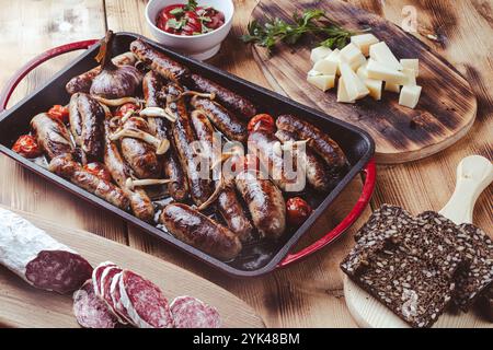 Grillpfanne mit leckeren hausgemachten Hühnchen-, Rind- und Schweinefleisch-Würstchen, Pilzen und Tomaten auf einem Holztisch mit Ketchup, Brot und Käse. Hoch Stockfoto