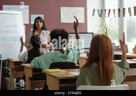 Rückansicht einer Reihe von cleveren interkulturellen Schülern, die Hände heben, während sie am Schreibtisch sitzen und dem Lehrer beim Unterricht zuhören Stockfoto