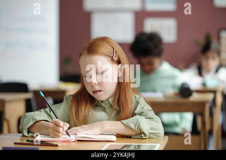 Kluges und fleißiges Schulmädchen, das Notizen auf der Seite des Copybook macht, während es am Schreibtisch im Klassenzimmer sitzt und Englischgrammatikunterricht erhält Stockfoto