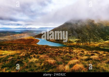 Das Foto zeigt einen See am Fuße einer Bergkette mit einem trockenen Gras im Vordergrund, einem großen, nebelbedeckten Hügel und einem Fernblick Stockfoto