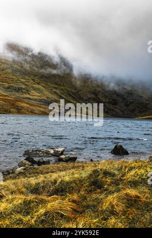 Ein ruhiger See spiegelt nebelige Hügel und Felsformationen wider, umgeben von üppigem Grasland. Diese malerische Umgebung fängt die Schönheit der Natur in einer Entfernung ein Stockfoto