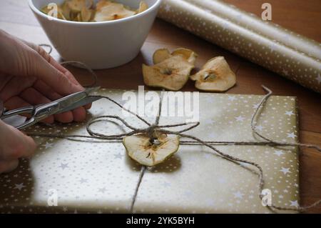 Nahaufnahme von weiblichen Händen, die Weihnachtsgeschenk umhüllen. Weihnachtsgeschenk in goldenem Geschenkpapier, verziert mit getrocknetem Apfel, getrockneten Äpfeln in einer Schüssel, Stockfoto