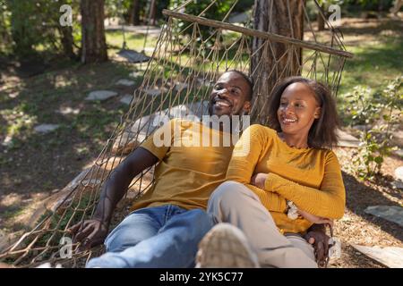 Sorglos lächelndes afroamerikanisches Paar, das seinen Urlaubstag in einer Hängematte verbringt und eine wunderschöne Glamping-Umgebung genießt. Stockfoto