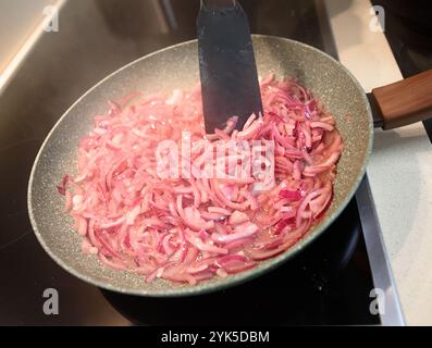 In dünnen Scheiben geschnittene rote Zwiebeln werden in einer Pfanne bei mittlerer Hitze sautiert und geben aromatische Düfte ab. Stockfoto