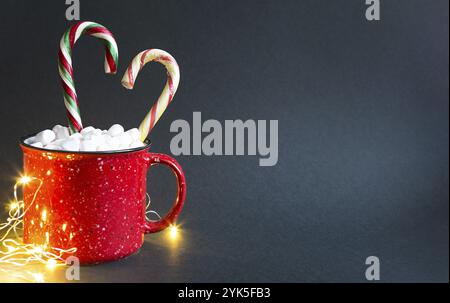 Roter Becher mit Marshmallows und Zuckerrohr in Herzform und Lichter von Girlanden auf schwarzem Hintergrund. Weihnachten, Neujahr, festliches Mo Stockfoto