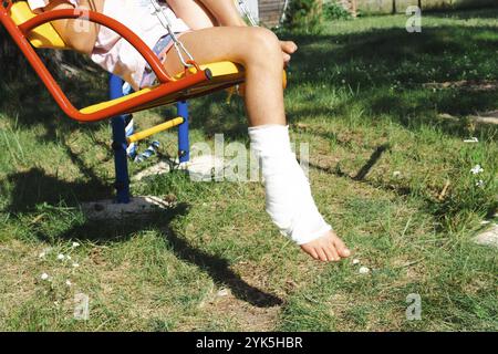 Kinderbeine sind mit Gips verbunden, barfuß auf einer Schaukel. Traumatische Ausrüstung, Kinderverletzung, Blutergüsse, Verstauchung, Fraktur Stockfoto