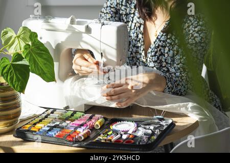 Eine Frau näht Tüll an eine elektrische Nähmaschine in einem weißen, modernen Innenraum eines Hauses mit großen Fenstern und Zimmerpflanzen. Komfort im Haus, ein Haus Stockfoto