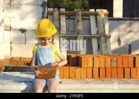 Das kleine Mädchen in einem gelben Hardhut spielt Baumeister auf der Baustelle ihres zukünftigen Heims. Erwartung, sich zu bewegen, einen Beruf zu wählen, Kinder Stockfoto