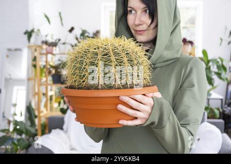 Grosser echinocactus Gruzoni in den Händen einer Frau im Inneren eines Grünhauses mit Regal-Sammlungen von Hauspflanzen. Eigenernte Stockfoto